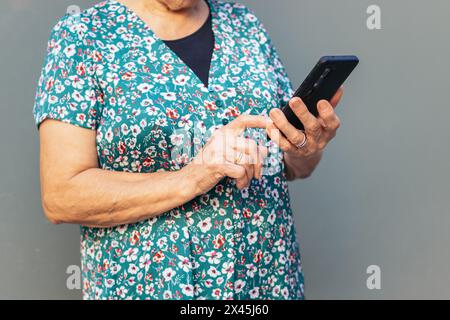 Primo piano di una donna anziana irriconoscibile con abiti colorati che utilizza lo smartphone appoggiato su una parete grigia Foto Stock