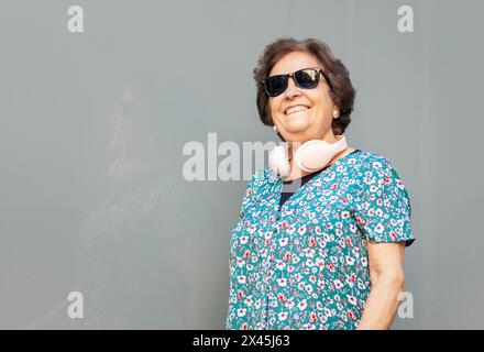 Ritratto di una donna anziana elegante con abiti colorati, occhiali da sole e cuffie rosa. Sfondo grigio. Foto Stock