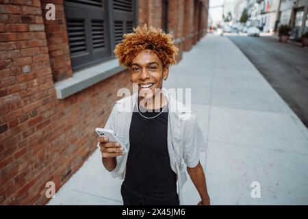 Un giovane uomo felice in outerwear e occhiali che cammina sul marciapiede, tenendo il cellulare Foto Stock