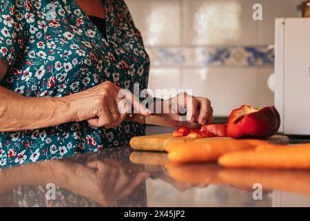 Primo piano di una donna anziana con bicchieri che preparano un pasto delizioso a casa. Taglia dei peperoni rossi in cucina Foto Stock