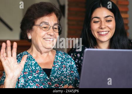 Una donna anziana sorridente che utilizza un laptop a casa e fa una videochiamata insieme a sua nipote. La nonna ondeggia alla telecamera Foto Stock
