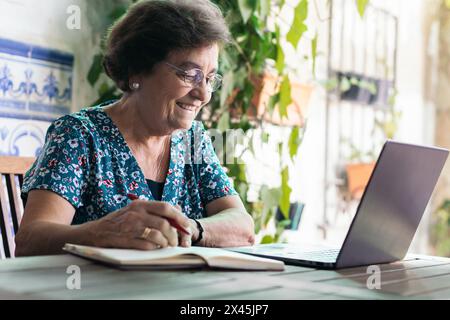 Immagine di una donna anziana che scrive su un notebook mentre usa il suo notebook a casa. Foto Stock
