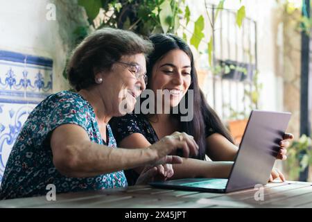 Donna anziana sorridente che usa un notebook. Accanto a lei, sua nipote la aiuta pazientemente a comprendere le nuove tecnologie. Foto Stock
