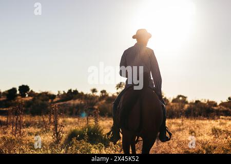 Uomo che indossa un cappello mentre cavalca un cavallo in campagna mentre il sole tramonta dietro di lui. Foto Stock