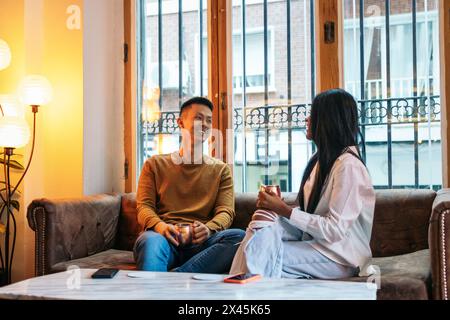 Uomo asiatico e donna latinoamericana seduti sul divano in uno spazio interno con luce calda. Parlano rilassati mentre bevono un drink. Foto Stock