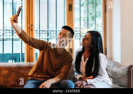 Amici multirazziali che scattano un selfie seduto su un divano in un interno con una grande finestra. Sorridono guardando la fotocamera dello smartphone Foto Stock