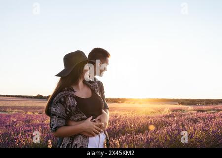 Splendida fotografia retroilluminata di una giovane coppia amorevole che passeggiava attraverso un campo di lavanda al tramonto. Foto Stock