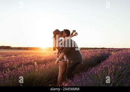 Una giovane coppia innamorata celebra il proprio anniversario con un appuntamento romantico in un campo di lavanda al tramonto. Il tizio prende la sua ragazza tra le braccia, a Foto Stock