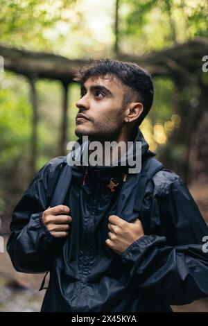 Giovane con impermeabile e zaino che cammina attraverso una giungla con grandi alberi, guardando in alto Foto Stock