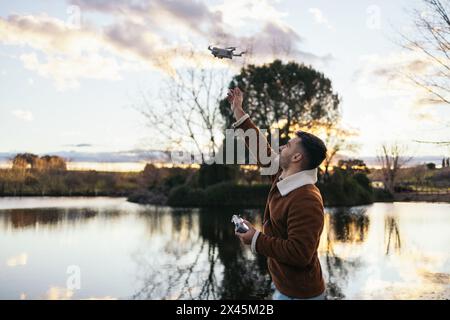 Giovane che usa un drone vicino a un lago per registrare e fotografare il paesaggio. Tiene il controller in una mano e con l'altra va al catc Foto Stock