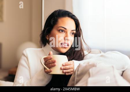 Giovane donna che si gode una tazza di caffè mentre si siede sul divano. Sembra che la donna si stia godendo il suo tempo libero Foto Stock