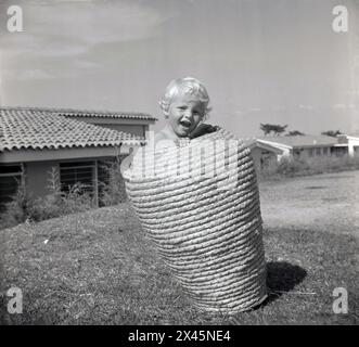 Anni '1960, storico, all'esterno su un'area di prato, un bambino dai capelli biondi in piedi in un alto cesto rotondo in vimini, Uganda. Foto Stock