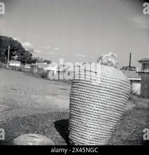 Anni '1960, storico, all'esterno su un'area di prato, visto da dietro, un bambino dai capelli biondi in piedi in un alto cesto rotondo di vimini, Uganda. Foto Stock