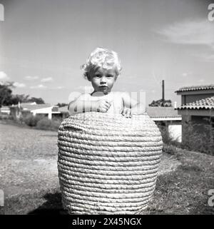 Anni '1960, storico, all'esterno su un'area di prato, un bambino dai capelli biondi in piedi in un alto cesto rotondo in vimini, Uganda. Foto Stock