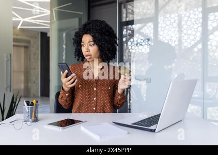 Una giovane donna nera scioccata alla scrivania del suo ufficio, che guarda con perplessità il suo smartphone mentre tiene in mano una carta di credito, probabilmente ha a che fare con una transazione inaspettata. Foto Stock