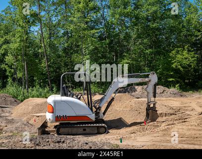 Un miniescavatore con paletta idraulica si trova sullo sporco in un nuovo lotto di costruzione della casa, con alberi sul retro del sito. Foto Stock