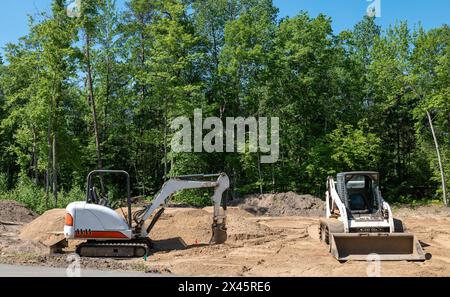 Un miniescavatore e un caricatore frontale compatto si trovano sullo sporco in un nuovo lotto di costruzione, con alberi sul retro del sito, in una giornata di sole con cielo blu. Foto Stock