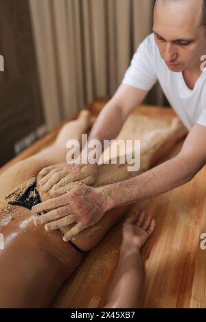 Shot verticale di massaggiatore maschile scrubbing massaggio natiche abbronzate con sabbia calda di una cliente donna sdraiata sul tavolo nel centro benessere. Foto Stock