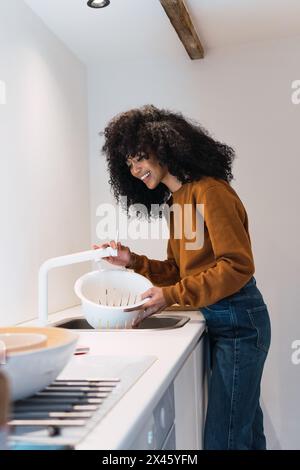 Ragazza allegra in caldo maglione e jeans che lavano ceci in colander nel lavandino mentre cucinano in cucina a casa Foto Stock