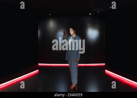 Vista laterale del corpo della giovane donna in elegante messaggio di testo abito sul cellulare mentre si trova in studio scuro con pareti nere Foto Stock