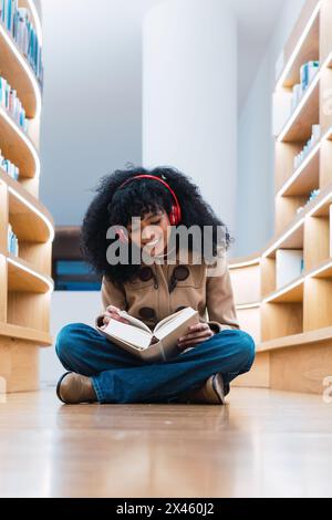 Felice studentessa afroamericana in cuffie leggere libro mentre si siede sul pavimento in biblioteca e godere Foto Stock