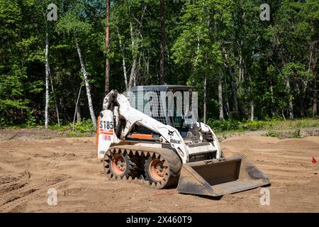 CROW WING CO, MN - 6 giugno 2021: Un caricatore frontale compatto Bobcat S185, bianco, con cingoli minipala gommata, si trova sullo sporco in un lotto di edifici vuoto fiancheggiato da alberi, Foto Stock