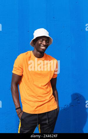 Giovane allegro maschio afroamericano in maglietta giallo brillante e cappuccio bianco in posa mentre si sta contro la parete blu Foto Stock