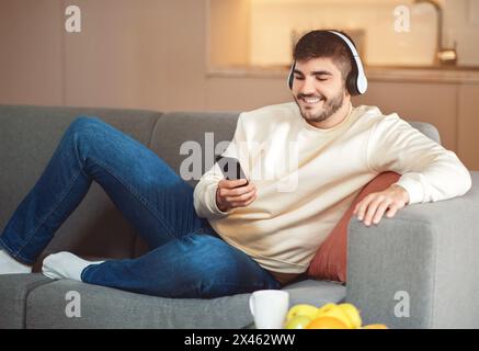 Giovane uomo che si rilassa sul divano indossando le cuffie e tenendo in mano lo smartphone Foto Stock