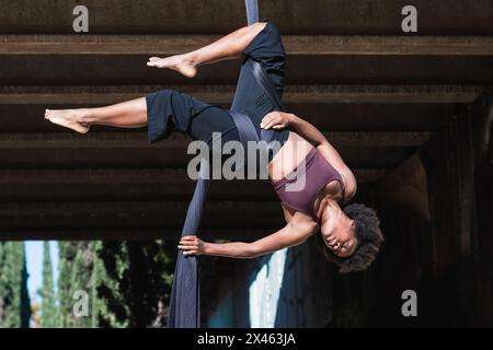 Bilanciamento femminile afroamericana capovolto su cinghie in fessure mentre si pratica lo yoga aereo in città Foto Stock