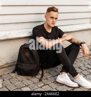 Giovane e affascinante ragazzo con Un abito nero alla moda e scarpe bianche seduti vicino a un muro di legno. Zaino nero. Foto Stock