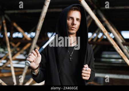 Angry Man in Black Clothes Holding A Bat sullo sfondo di Metal Pipes Foto Stock