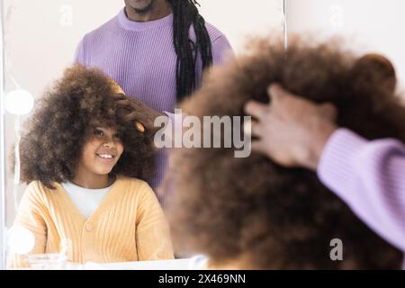 Raccolto africano americano padre che combatte i capelli ricci di ragazza seduta vicino allo specchio e sorridendo dolcemente Foto Stock