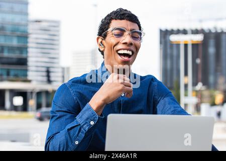 Adulto etnico allegro maschio remoto dipendente in auricolare che lavora su netbook in città il giorno di sole Foto Stock