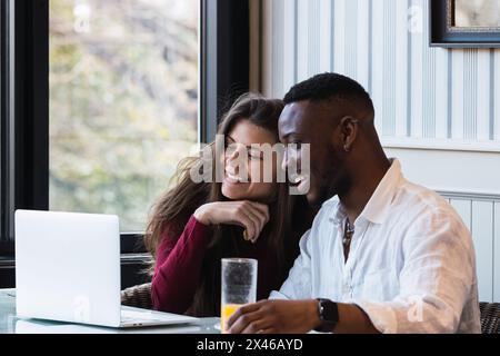 Felice coppia multirazziale seduto al tavolo e guardando il laptop insieme mentre si ha la colazione in una camera elegante a casa Foto Stock