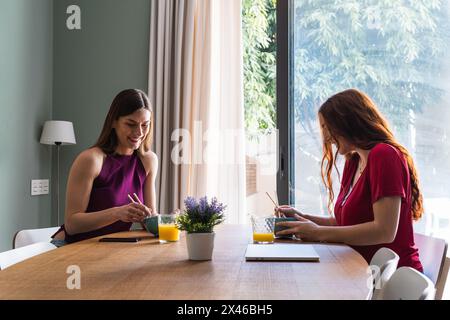 Felice giovane femmina amici in abiti casual avendo pranzo delizioso insieme e chiacchierando mentre si riuniscono al tavolo da pranzo in luce appartamento Foto Stock