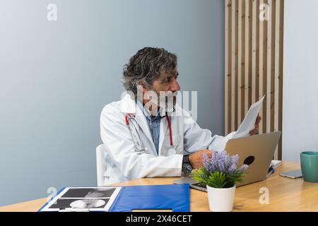 Anziano medico maschile seduto al tavolo in studio medico e l'esame dei raggi X mentre si lavora in clinica Foto Stock