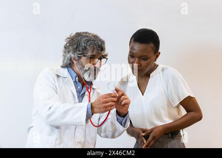 Medico maschile senior focalizzato che mostra stetoscopio alla paziente femminile nera durante il check-up in ospedale Foto Stock