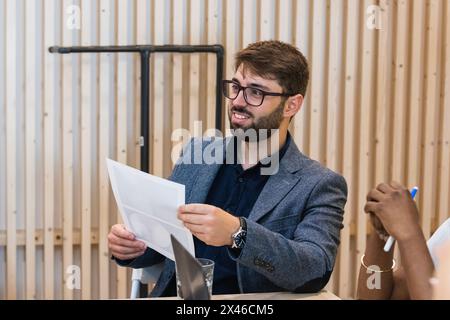 Allegro imprenditore maschile seduto sul posto di lavoro con Crop black collega e l'esame di relazione durante il lavoro sul progetto Foto Stock