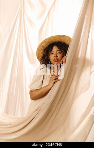 Una bella giovane donna afroamericana con capelli ricci in un cappello di paglia che sbircia fuori da una tenda in un ambiente da studio. Foto Stock