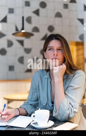 Giovane donna pia che si appoggia a portata di mano al tavolo con una tazza di bevanda calda e il blocco note mentre si guarda lontano in caffetteria Foto Stock