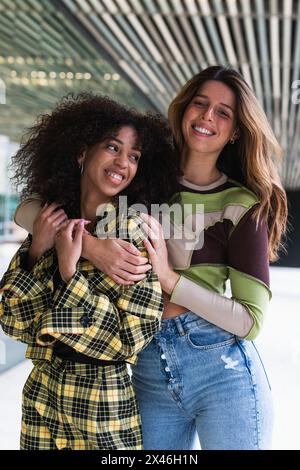 Giovane donna sorridente che abbraccia allegra ragazza etnica con acconciatura afro in abito a scacchi alla moda in città Foto Stock