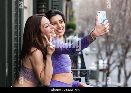 Vista laterale di felice giovane donna ispanica sorridente con un amico che fa gesto pezzo mentre si prende selfie su smartphone in piedi sul balcone Foto Stock