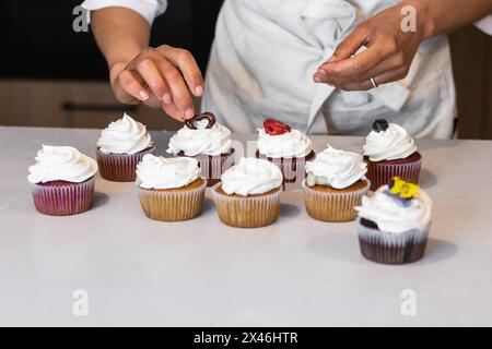 Panettiere femminile corto irriconoscibile in piedi al banco in cucina di panetteria e decorare dolci cupcake vegan con bacche Foto Stock