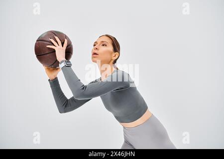 Una giovane donna sportiva tiene con grazia un pallacanestro con la mano destra su uno sfondo grigio neutro. Foto Stock