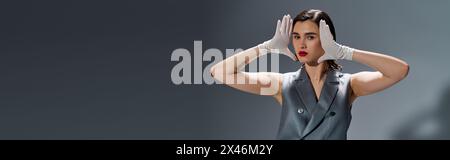 Una giovane donna elegante indossa un abito elegante con un giubbotto, le mani sulla testa, esprimendo emozioni in un ambiente da studio su uno sfondo grigio. Foto Stock