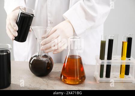 Donna che versa olio grezzo nero dal becher al matraccio sul tavolo grigio su sfondo chiaro, primo piano Foto Stock