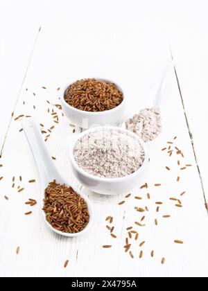 Farina di riso rossa nel recipiente e nel cucchiaio, cereali nel cucchiaio, nel recipiente e sul tavolo sullo sfondo di un bordo di legno bianco Foto Stock