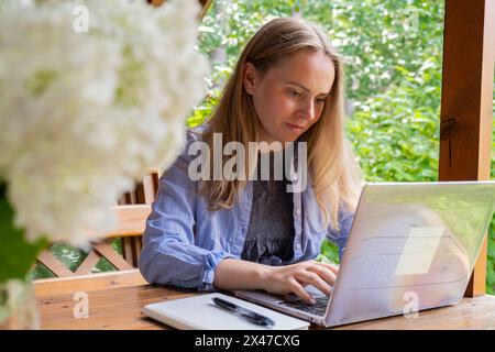 La giovane donna felice si concentra sul suo notebook in un'alcova di legno. L'ambiente esterno rilassato enfatizza comfort e produttività. Concetto di apprendimento del lavoro a distanza Foto Stock