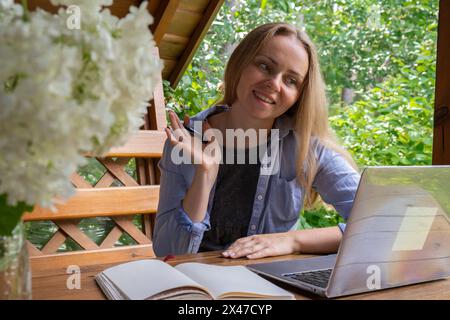 Giovane lavoratrice freelance che lavora online utilizzando un computer portatile e si gode la splendida natura all'aperto in giardino. Formazione per riunioni online. Worcation, lavoro dalle vacanze, modello di lavoro ibrido Foto Stock