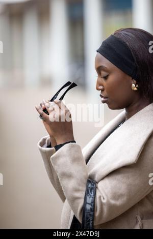 Una donna afroamericana viene raffigurata mentre esamina un paio di occhiali da sole all'aperto. È vestita con un cappotto beige leggero con cintura nera e fascia, trasuda un senso di casual urban fashion. La bassa profondità di campo mette l'attenzione su di lei, con dettagli architettonici leggermente sfocati sullo sfondo. La sua espressione è concentrata e serena. Donna afroamericana che controlla gli occhiali da sole su Urban Street. Foto di alta qualità Foto Stock